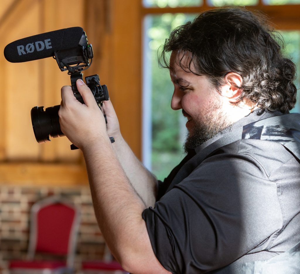Image of Tom holding a camera with a microphone on top of it and smiling.